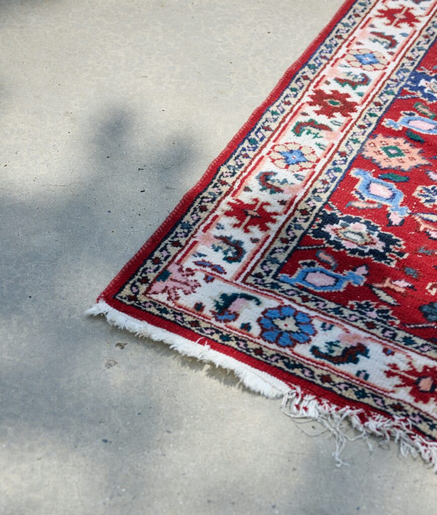 Fringed corner of a red oriental rug with floral patterns on concrete surface, ideal for professional rug cleaning services