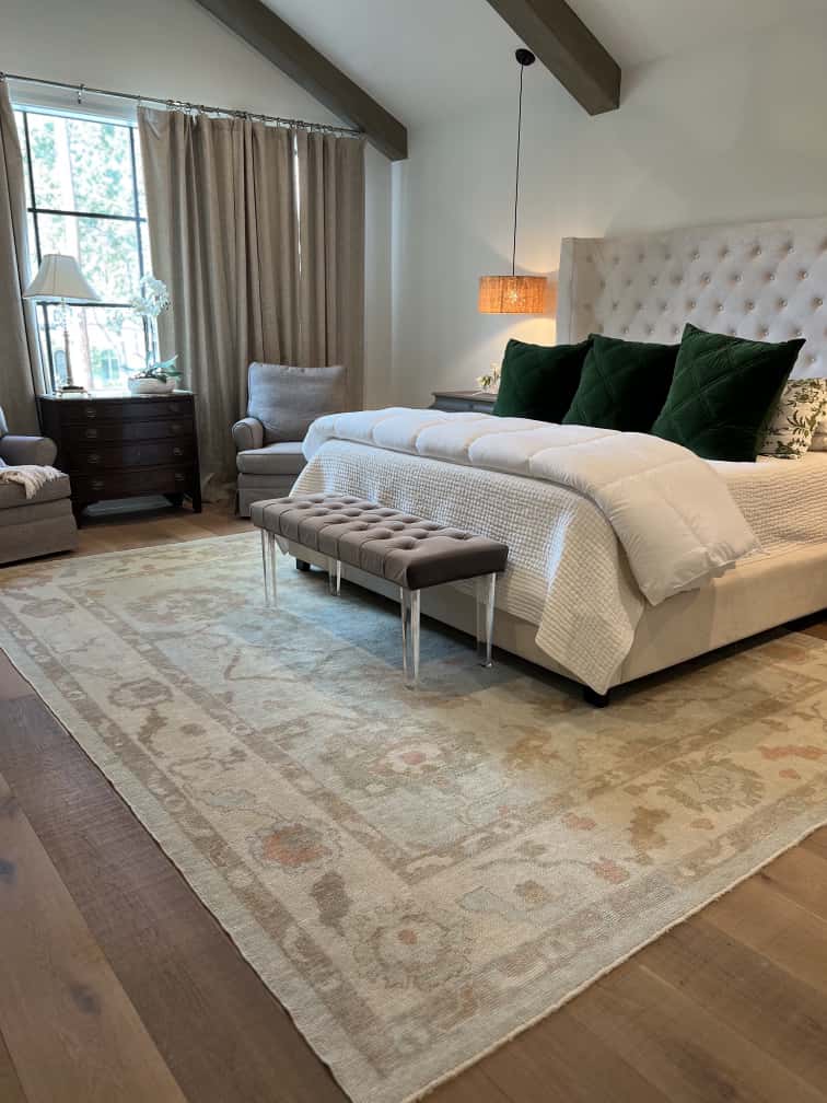 Elegant bedroom interior featuring a soft neutral-toned Turkish Oushak rug with subtle floral patterns. The rug complements the tufted headboard, green velvet pillows, and natural wood flooring, creating a calm and inviting atmosphere. Available exclusively at RugGuys in Dallas–Fort Worth, specializing in authentic handmade Oushak rugs.
