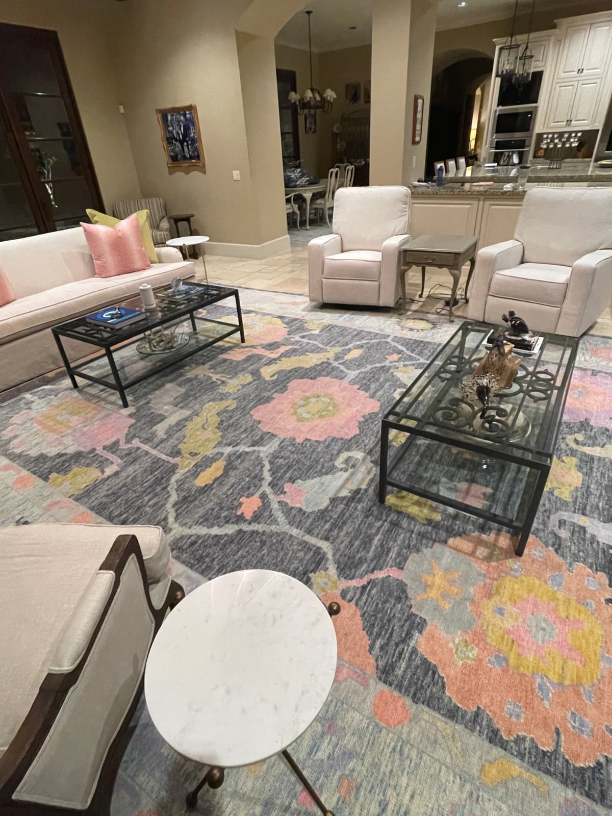 Contemporary living room featuring a colorful handmade Turkish Oushak rug with floral patterns in pink, yellow, and gray tones. The soft wool rug adds warmth and personality to the neutral seating area with beige walls and glass-top coffee tables. Available at RugGuys in Dallas–Fort Worth, specializing in luxury handmade rugs.