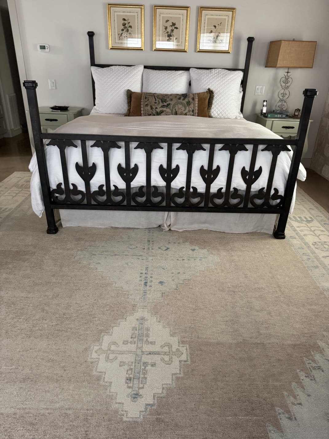Elegant bedroom featuring a neutral-toned Turkish Oushak rug with soft tribal medallion motifs, paired with a black four-poster bed, white bedding, botanical wall art, and classic décor.
