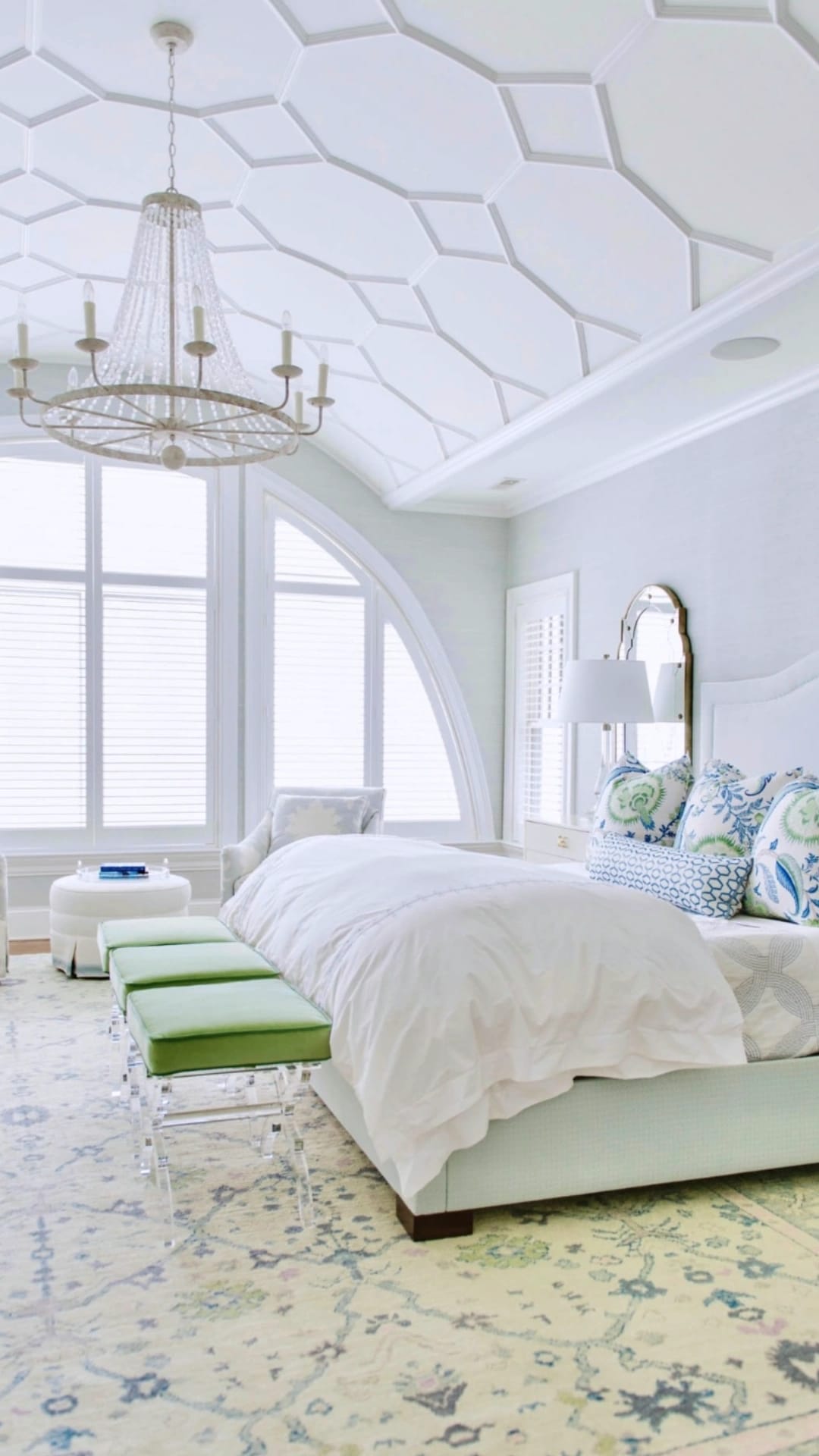 Bright, elegant bedroom featuring a soft pastel Turkish Oushak rug with floral motifs, styled with white bedding, green bench seating, large arched windows, and a decorative chandelier.