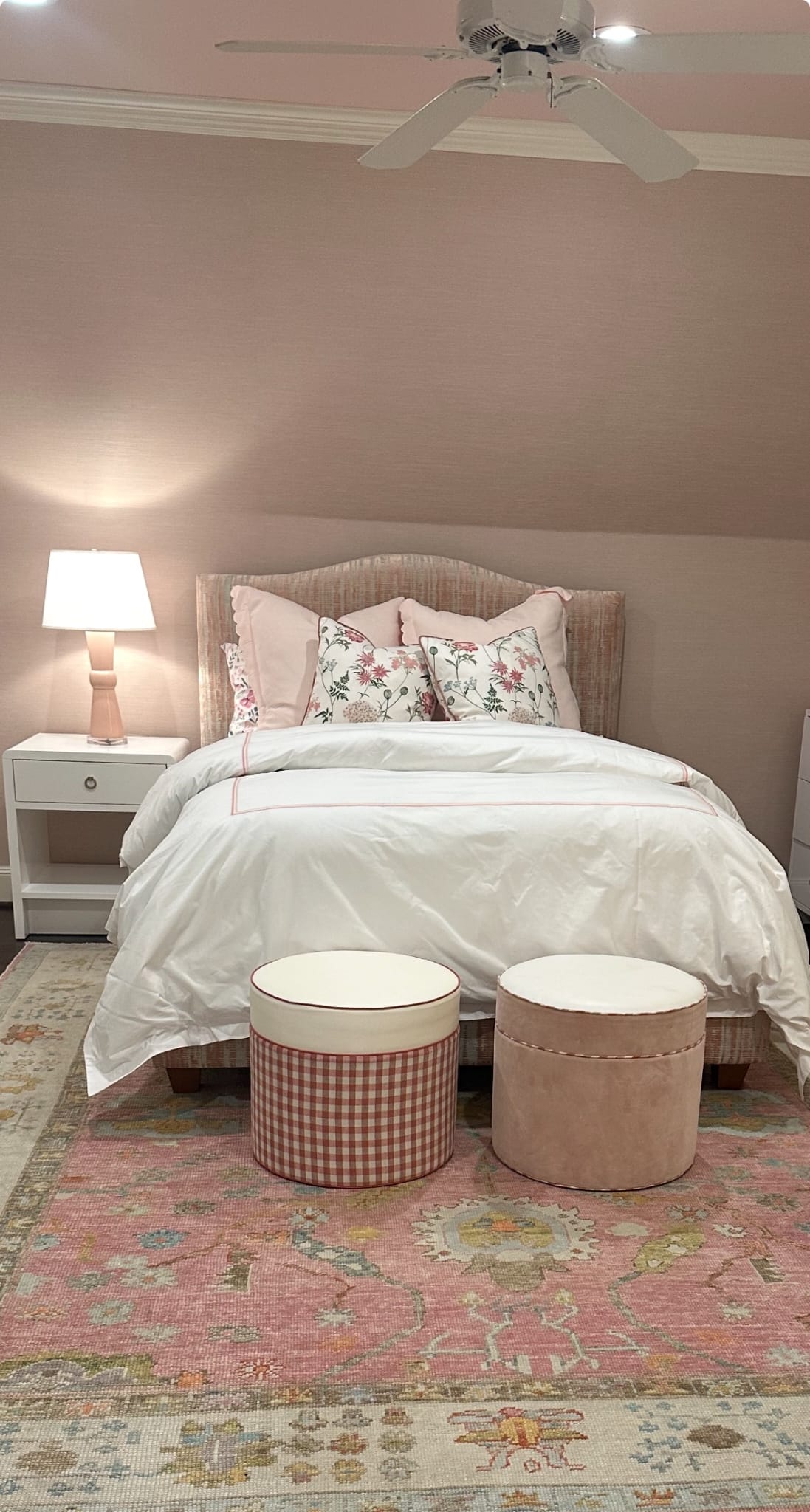 Beautiful bedroom styled with a soft pink Turkish Oushak rug featuring pastel floral motifs, paired with white bedding, floral pillows, and gingham and velvet ottomans for a cozy, feminine look.