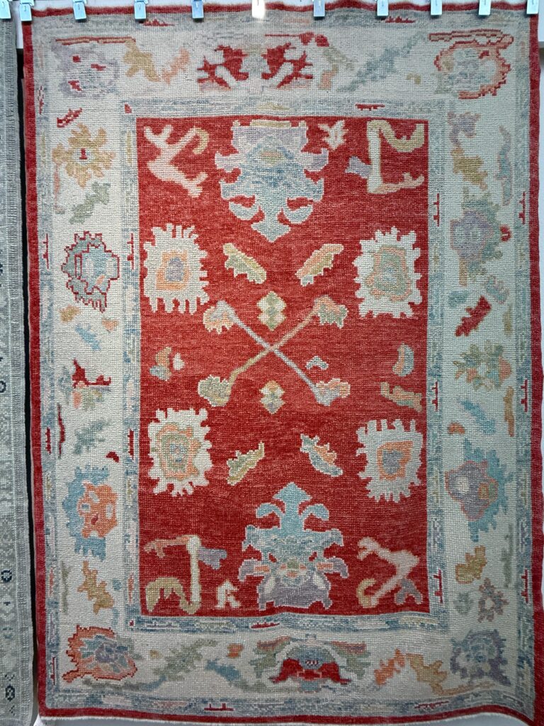 Handwoven Turkish Oushak rug featuring a bold red center field, surrounded by a soft muted border with pastel tribal and floral motifs in blue, cream, gold, and peach. Vintage-inspired wool texture with traditional geometric patterns, available at RugGuys Dallas for upscale interior design.