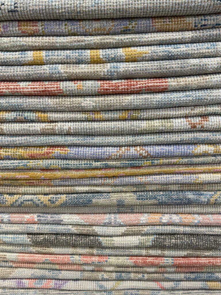 Stack of authentic handwoven Turkish Oushak rugs folded neatly, showcasing soft pastel tones, vintage-inspired motifs, and traditional wool textures. Premium handmade rugs available at RugGuys in Dallas, perfect for living rooms, bedrooms, and designer interiors.